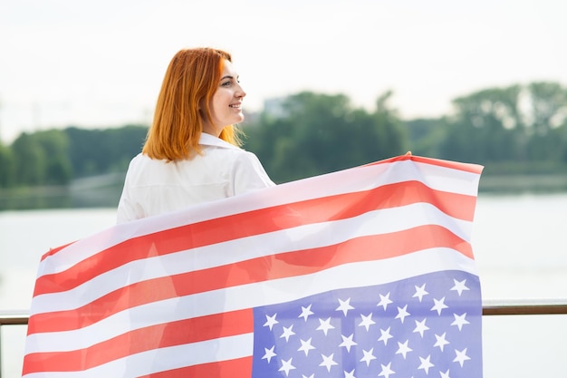 Visão traseira de uma jovem feliz com a bandeira nacional dos EUA nos ombros Garota positiva comemorando o dia da independência dos Estados Unidos