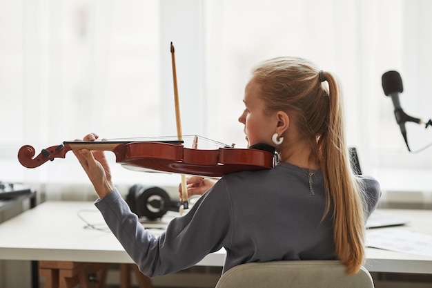 Visão traseira de uma jovem loira tocando violino no estúdio contra o espaço de cópia de luz da janela