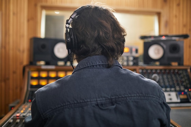 Visão traseira do homem usando fones de ouvido na estação de trabalho de áudio no espaço de cópia de estúdio de gravação profissional