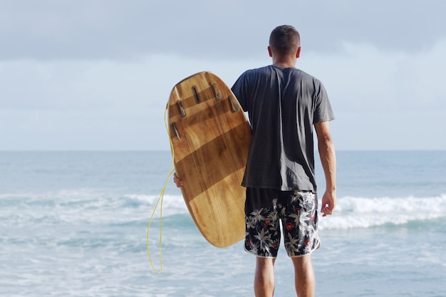 Visão traseira. Surfista com prancha de surf, olhando para o oceano de manhã.