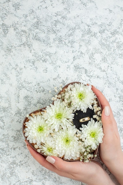 Visão vertical da mão segurando uma linda flor e anel em caixa em forma de coração em fundo branco gelado