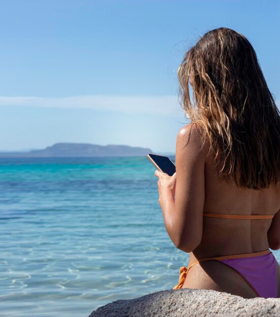 Visão vertical da mulher irreconhecível usando smartphone na praia com água turquesa clara