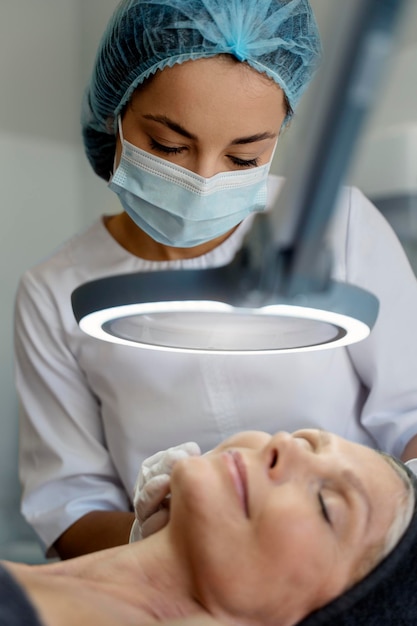Visão vertical de uma jovem massagista massageando o rosto de uma paciente sênior sob uma lâmpada especial
