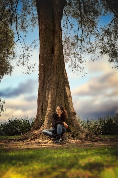 Foto visão vertical onde uma jovem é mostrada em primeiro plano sentada à sombra de uma árvore muito grande e ao fundo um céu muito bonito