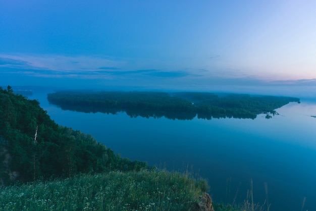 Visión mística de la orilla alta en el río ancho con agua suave. Orilla del río de la gran isla con bosque bajo la niebla. Neblina temprana sobre los árboles.