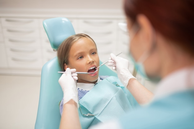 Visita al dentista infantil. Hermosa colegiala de pelo oscuro visitando al dentista infantil en la mañana