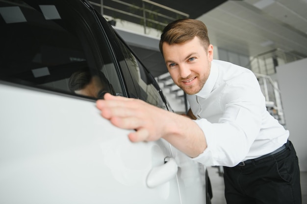 Visitando el concesionario de automóviles Un hombre guapo con barba está acariciando su auto nuevo y sonriendo