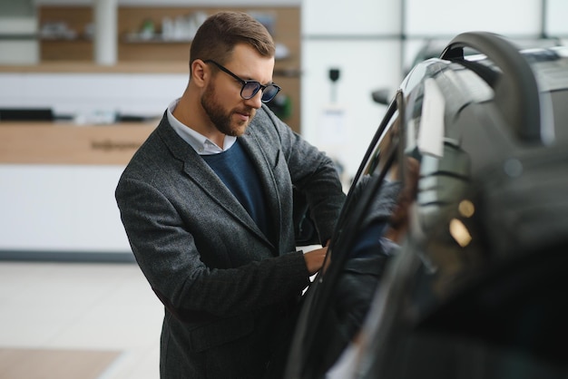 Visitando el concesionario de automóviles Un hombre guapo con barba está acariciando su auto nuevo y sonriendo