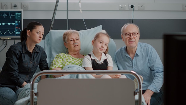 Visitantes viendo la televisión con un paciente anciano en la cama de la sala de hospital. Madre y niña visitando a un pensionista enfermo, mirando la televisión y disfrutando de la visita con la familia. Personas que apoyan a la mujer