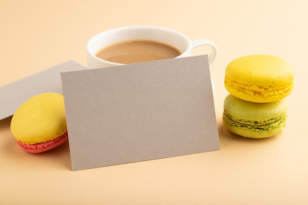 Visitenkartenmodell aus grauem Papier mit gelben und grünen Makronen und einer Tasse Kaffee auf orangefarbenem Pastellhintergrund, Seitenansicht, Kopienraum