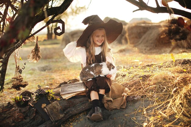 Víspera de Todos los Santos. alegre bruja con una varita mágica y un libro conjura y se ríe. niña en un traje de bruja niña sosteniendo un conejo en sus brazos