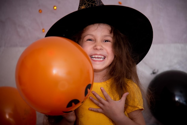 Víspera de Todos los Santos de los niños. Una niña sonriente con un sombrero de bruja y una camiseta naranja con un frasco de vidrio en forma de calabaza se prepara para celebrar.