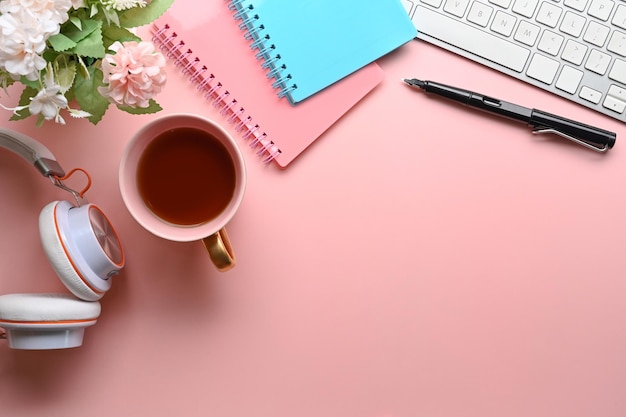 Vista acima do espaço de trabalho da mulher com teclado de notebook de xícara de café de fone de ouvido e flores em fundo rosa pastel