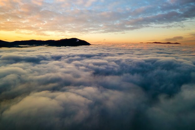 Vista aérea del amanecer vibrante sobre densas nubes blancas con montañas oscuras distantes en el horizonte.