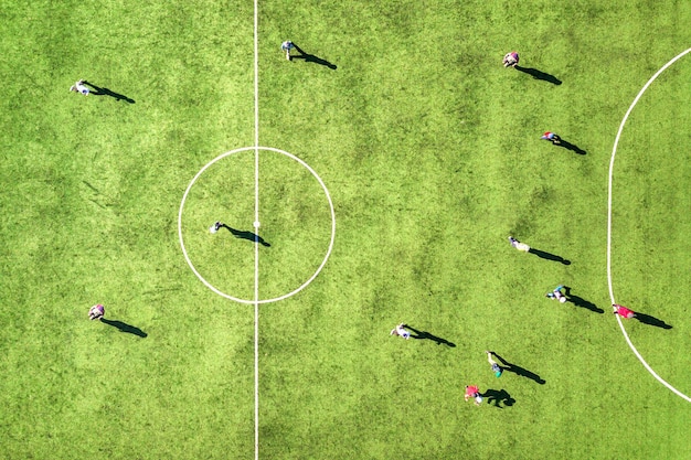 Vista aérea de arriba hacia abajo del campo de deportes de fútbol verde y jugadores jugando al fútbol. Drone tomó la imagen de pequeños deportistas irreconocibles en el estadio cubierto de hierba durante las actividades deportivas.