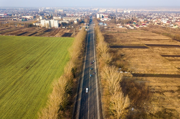 Vista aérea de la carretera con coches en movimiento, campos verdes y arados y praderas y suburbios de la ciudad en un día soleado. Fotografía de drones.