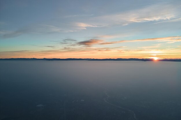 Vista aérea de la colorida puesta de sol sobre densas nubes de niebla blancas cubiertas con siluetas oscuras distantes de colinas de montaña en el horizonte