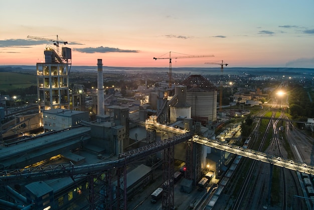 Vista aérea da fábrica de cimento com alta estrutura de planta de concreto e guindastes de torre na área de produção industrial à noite. Fabricação e conceito de indústria global.