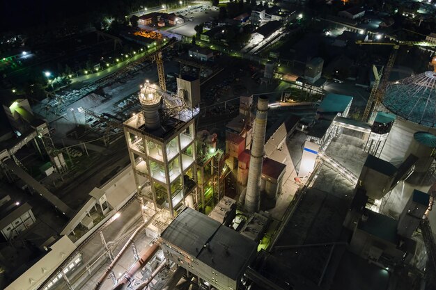 Vista aérea da fábrica de cimento com alta estrutura de planta de concreto e guindastes de torre na área de produção industrial à noite. Fabricação e conceito de indústria global.