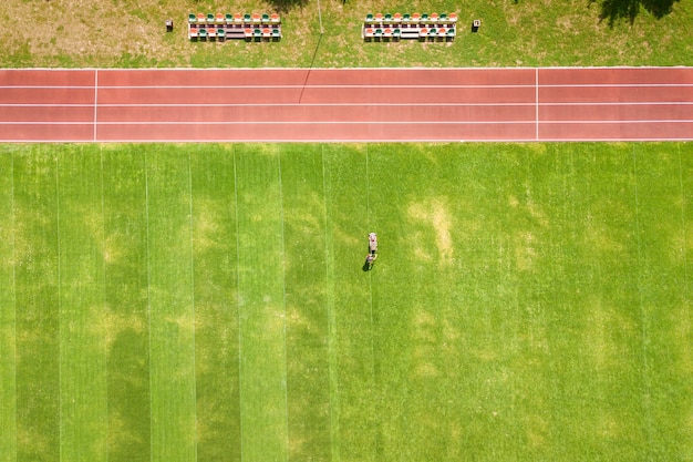 Vista aérea da pequena figura de trabalhador cortando a grama verde com mashine no campo do estádio de futebol com pistas de corrida vermelhas no verão.