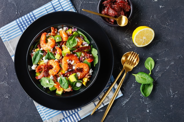 Vista aérea da salada de espinafre com abacate e camarão com tomates secos ao sol, pedaços de bacon frito, queijo esfarelado, cebola roxa em uma tigela preta sobre uma mesa de concreto