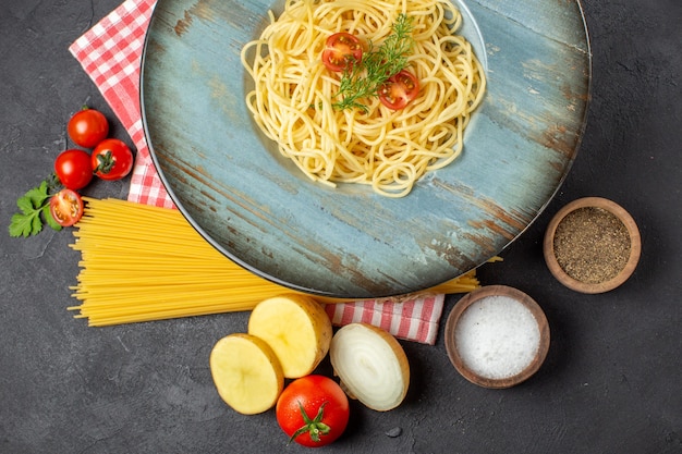 Vista aérea de um delicioso spagetti servido com tomates verdes e diferentes especiarias massas crus em uma toalha vermelha despojada na mesa preta