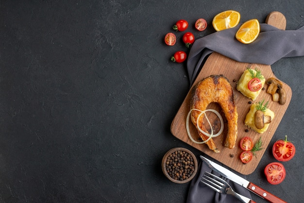 Vista aérea de uma deliciosa refeição de peixe frito com cogumelos, tomate, queijo na placa de madeira, fatias de limão, pimenta em talheres de toalha de cor escura definir pimenta do lado esquerdo na superfície preta angustiada