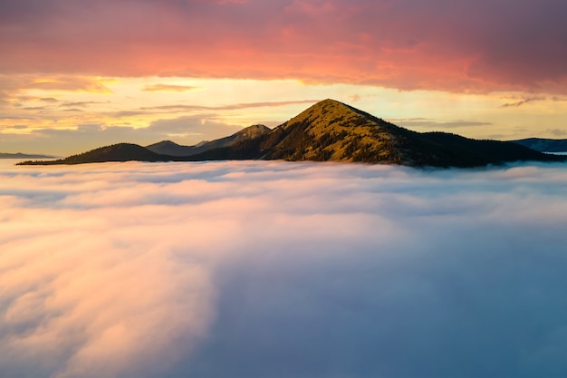 Vista aérea de uma grande montanha sobre nuvens brancas densas ao amanhecer.