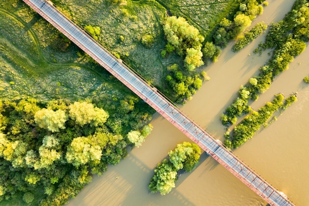 Vista aérea de uma ponte de estrada estreita que se estende ao longo de um rio lamacento na área rural verde.