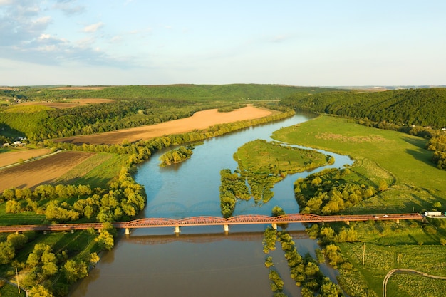 Vista aérea de uma ponte de estrada estreita que se estende ao longo de um rio lamacento na área rural verde.