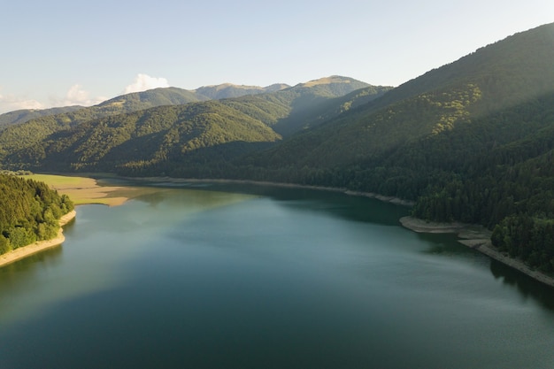 Vista aérea do grande lago com água azul clara entre colinas de alta montanha cobertas por densa floresta perene.