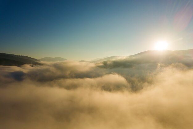 Vista aérea do nascer do sol vibrante sobre a névoa branca e densa com silhuetas escuras distantes das colinas de montanha no horizonte.