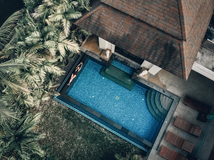 Vista aérea do território do hotel com palmeiras, gramado verde, telhado de telhas e a piscina clara com uma garota magra flutuando nas costas; conceito de luxo.