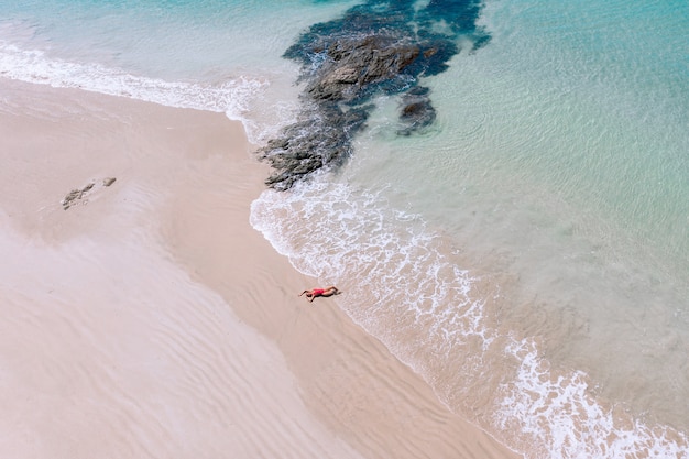 Vista aérea de drone de mujer joven en bikini rojo tomar el sol en la playa con arena blanca