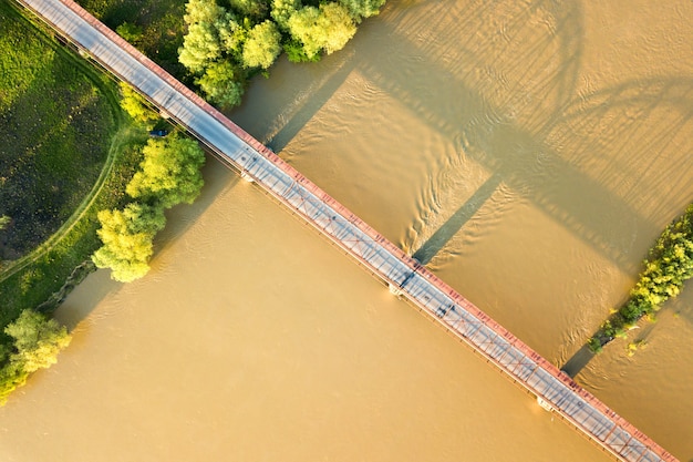 Vista aérea de un estrecho puente de carretera que se extiende sobre ancho río fangoso en zona rural verde.