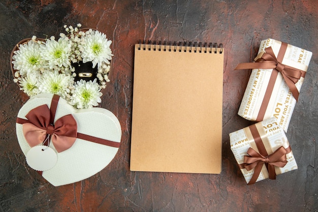 Vista aérea de hermosas flores en caja en forma de corazón con anillo y cuaderno de regalos sobre fondo de color mixto con espacio libre