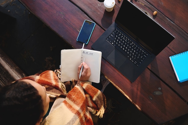 Vista aérea de una mujer escribiendo en un cuaderno mientras está sentado en un banco de madera frente a una computadora portátil y suministros de oficina. Concepto de e-learning, trabajo remoto, negocio remoto durante el bloqueo de cuarentena