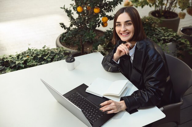 Vista aérea de una mujer independiente, joven empresaria, redactora, sonriendo tiernamente mirando a la cámara mientras está sentada a la mesa con un bloc de notas y una computadora portátil, trabajando en línea de forma remota en una terraza de la cafetería