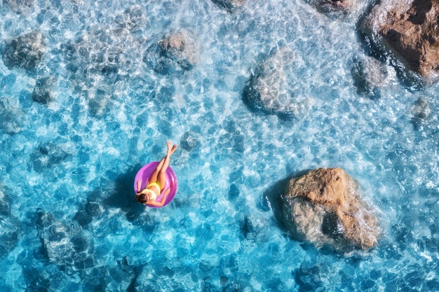 Foto vista aérea de una mujer joven nadando con anillo de natación rosa en el mar azul al atardecer en verano paisaje tropical con niña agua clara piedras playa de arena vista superior vacaciones en la isla de lefkada grecia