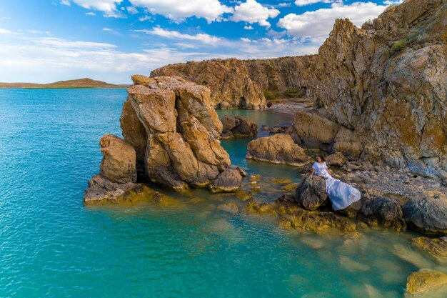 Vista aérea de una mujer joven con un vestido blanco en las rocas. Paisaje marino de verano con una niña, playa, hermosas olas, rocas, agua azul. Vista desde arriba. Naturaleza