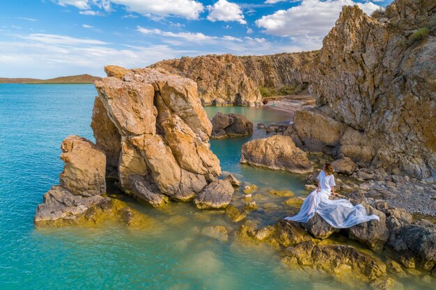 Vista aérea de una mujer joven con un vestido blanco en las rocas. Paisaje marino de verano con una niña, playa, hermosas olas, rocas, agua azul. Vista desde arriba. Naturaleza