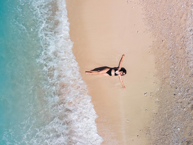 Vista aérea de la mujer en traje de baño negro tomando el sol en la playa de arena de la orilla del mar