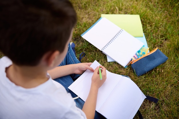 Vista aérea del niño de la escuela escribiendo en hojas en blanco vacías de un libro. Vista trasera de un niño de primaria haciendo tarea escolar en el parque después de la escuela. Accesorios escolares recostados sobre el césped