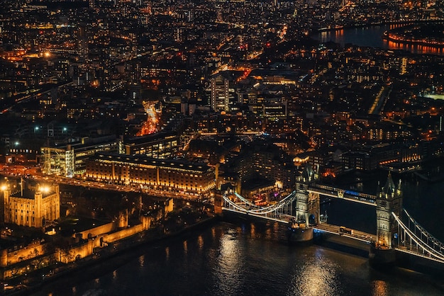 Vista aérea noturna da Tower Bridge sobre o rio Tâmisa, edifícios iluminados brilhando no escuro