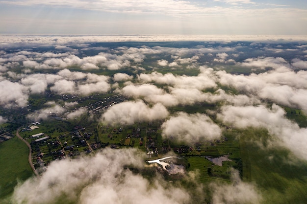 Vista aérea de nubes blancas sobre una ciudad o pueblo con hileras de edificios y calles con curvas entre campos verdes en verano. Paisaje de campo desde arriba.