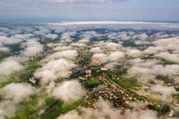 Vista aérea de nubes blancas sobre una ciudad o pueblo con hileras de edificios y calles con curvas entre campos verdes en verano. Paisaje de campo desde arriba.