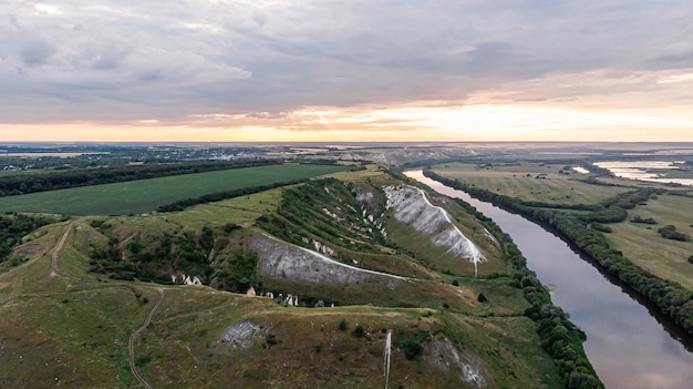 Foto vista aérea panorámica sobre la cima de un paisaje de verano de un campo agrícola verde, colinas o montañas, un gran río y un cinturón forestal al atardecer