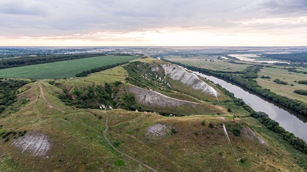 Vista aérea panorâmica sobre o topo de uma paisagem de verão de um campo agrícola verde, colinas ou montanhas, um grande rio e um cinturão de floresta ao pôr do sol