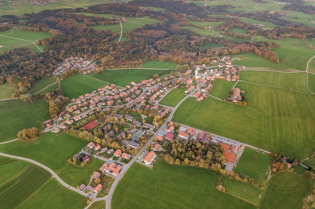 Vista aérea de la pequeña ciudad con techos de tejas rojas entre campos de cultivo verde y bosque distante en verano.