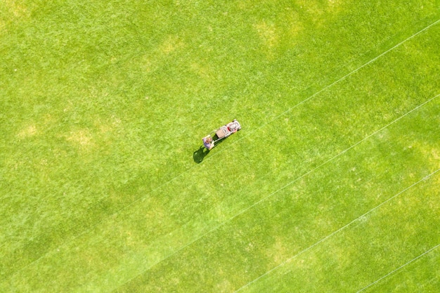 Vista aérea de la pequeña figura de trabajador hombre recortando pasto verde con mashine de corte en el campo del estadio de fútbol en verano.